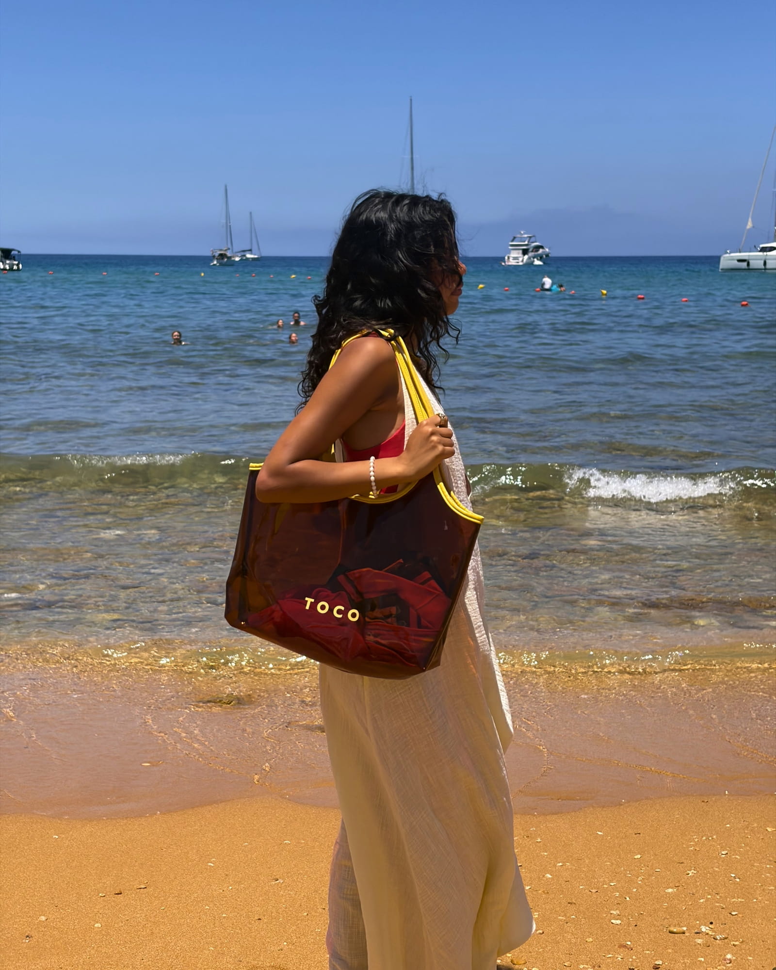 Woman in a white beach dress carrying the Toco amber jelly beach tote at the shoreline