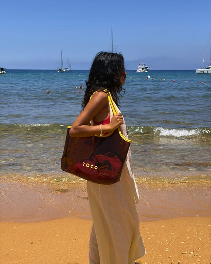 Woman in a white beach dress carrying the Toco amber jelly beach tote at the shoreline