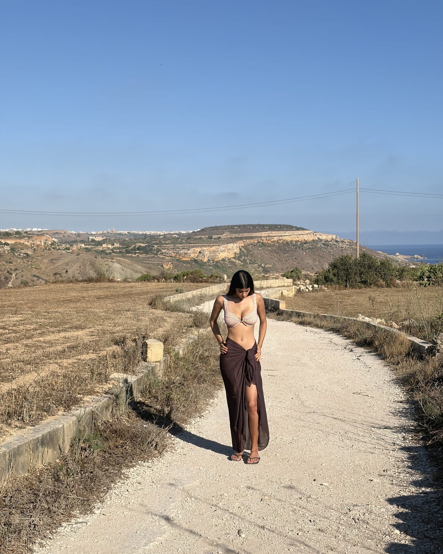 Woman on a countryside path wearing the Toco Swim Chocolate maxi sarong in solid brown, tied at the waist over a crochet bikini