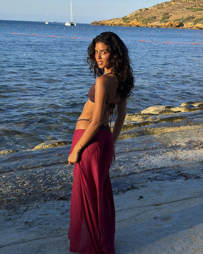 Back view of the Toco Swim Shiraz wine-burgundy maxi sarong tied at the hip, sea and boats behind at sunset
