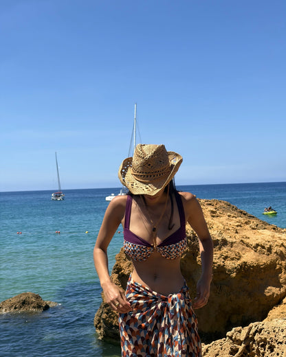 Woman on sunlit rocks by a turquoise sea wearing the Toco Swim Siren maxi sarong and matching bikini with a straw hat